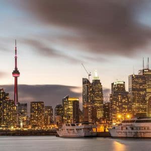 A city skyline filled with illuminated skyscrapers and a tall, red-lit tower at dusk, viewed from across the water where two boats are docked.