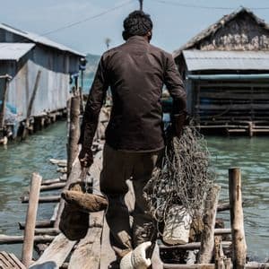 Un homme de dos à l'appareil photo transporte du matériel de pêche sur une jetée en bois dans un village sur pilotis.
