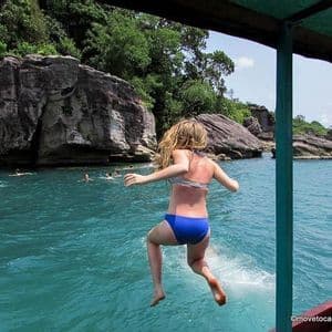 Une femme en bikini bleu saute d'un bateau dans l'eau turquoise, avec des falaises rocheuses et d'autres personnes qui nagent en arrière-plan.