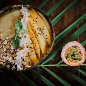 Un bol de smoothie mangue, granola et copeaux de coco dans une coque de noix de coco, près d'un fruit de la passion sur une feuille de palmier.