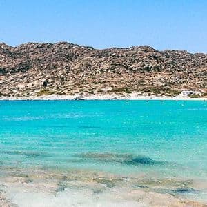Vista panoramica di una baia con acqua cristallina color turchese, una spiaggia di sabbia e una catena montuosa rocciosa sotto un cielo azzurro splendente.