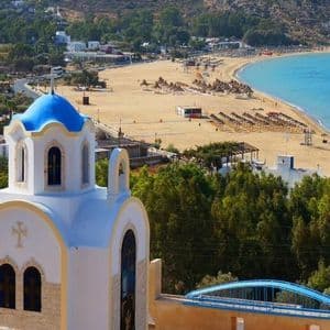 Una chiesa bianca con cupola blu si affaccia su un'ampia spiaggia sabbiosa con acqua turchese e file di ombrelloni.