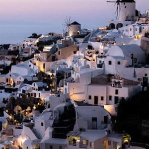 Edifici bianchi e mulini a vento raggruppati su una collina a picco sul mare, illuminati da luci calde al tramonto.