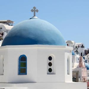 Una chiesa bianca con una cupola blu brillante e una croce in cima, incastonata tra un gruppo di edifici imbiancati a calce sotto un cielo sereno.