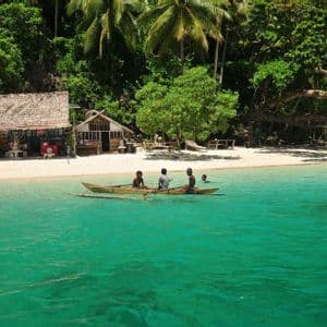 Tre persone remano una piccola canoa in acqua turchese al largo di una spiaggia di sabbia bianca con capanne di paglia e una giungla fitta.