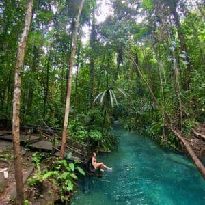 Una donna in bikini siede su una piattaforma di legno, immergendo i piedi in un limpido fiume turchese che attraversa una giungla fitta.