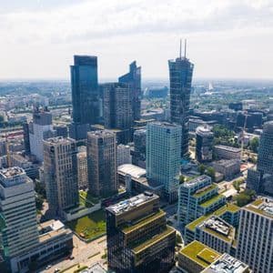 An aerial view of a modern city with glass skyscrapers and green-roofed buildings under a partly cloudy sky.