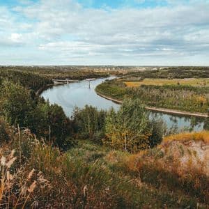 A high-angle view of a wide river curving through a lush, tree-filled valley with a distant bridge under a cloudy blue sky.