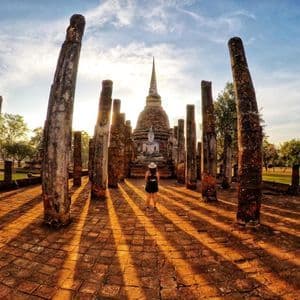 Eine Person steht inmitten antiker Säulen und blickt auf eine Buddhastatue und einen Stupa, während die untergehende Sonne lange Schatten wirft.