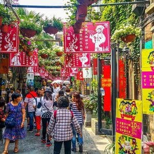Una folla di persone cammina lungo un vicolo stretto e vivace, decorato con striscioni rossi appesi e piante verdi.