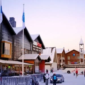 Menschen in Winterkleidung gehen in der Dämmerung eine schneebedeckte Straße entlang, die von Holzhäusern und Geschäften gesäumt ist.