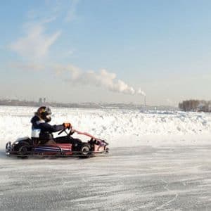 Eine Person mit Helm fährt ein Go-Kart auf einer gefrorenen, vereisten Strecke in einer verschneiten Landschaft mit einer Stadt in der Ferne.