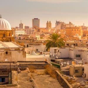 Vista sul paesaggio urbano dai tetti piatti, con un edificio dalla cupola bianca, una palma e moderni grattacieli nello sfondo nebbioso.