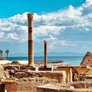 D'anciennes ruines de pierre avec de hautes colonnes patinées se dressent au bord de la mer sous un ciel bleu partiellement nuageux.
