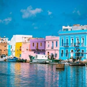 Bateaux amarrés dans un port devant une rangée de bâtiments aux couleurs vives sous un ciel bleu clair.