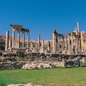 D'anciennes ruines de pierre, avec des rangées de colonnes, se dressent sur un champ herbeux sous un ciel bleu clair.