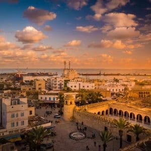 Vue aérienne d'une ville côtière historique, avec une place animée, un port et des navires, sous un spectaculaire coucher de soleil nuageux.