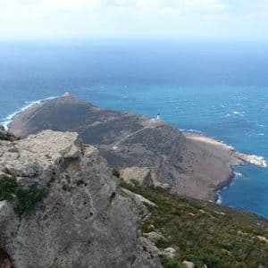 Vue d'une falaise rocheuse dominant une péninsule avec un phare blanc, entourée par la mer bleue.