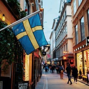 A Swedish flag hangs from a pole on a building along a narrow cobblestone street filled with shops and people.