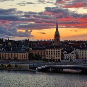 Un paesaggio urbano con un prominente campanile di chiesa si erge sulla riva di un fiume, sotto un cielo al tramonto dai colori vivaci, con nuvole arancioni e viola.