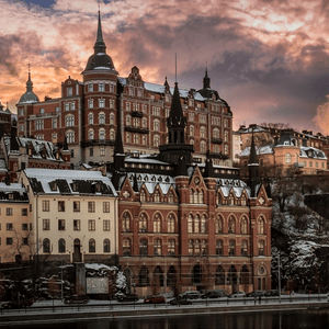 Ornate city buildings with snow-covered roofs stand on a rocky hillside under a dramatic sunset sky.