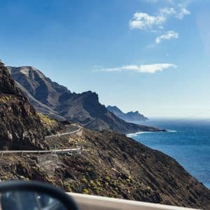 Una vista dall'auto di una strada tortuosa lungo una costa ripida e rocciosa accanto al mare blu profondo sotto un cielo soleggiato.