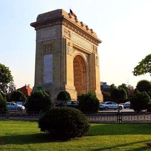 Un gran arco de triunfo de piedra se alza tras un verde césped de parque, con coches circulando por una carretera intermedia bajo un cielo despejado.
