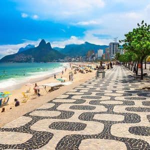 Ein sonniger, belebter Stadtstrand mit einer markanten schwarz-weiß gemusterten Promenade und hohen Bergen in der Ferne.