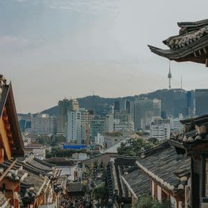 Una vista dall'alto di una strada affollata, fiancheggiata da case tradizionali con tetti di tegole, con uno skyline moderno della città sullo sfondo.