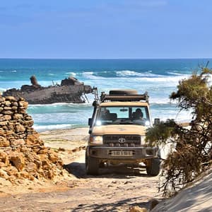 Un veicolo fuoristrada percorre un sentiero sabbioso verso una spiaggia con un relitto arenato tra le onde.