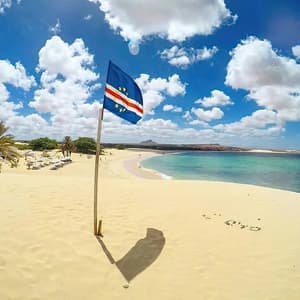 La bandiera di Capo Verde è piantata su una duna di sabbia che si affaccia su un'ampia spiaggia con acqua turchese sotto un cielo blu e nuvoloso.