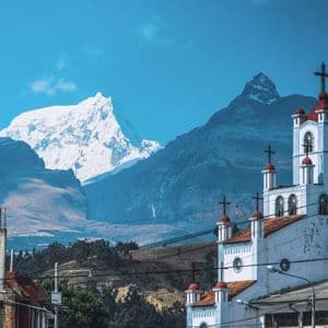 Una chiesa bianca dal tetto rosso sorge in un paese ai piedi di una grande catena montuosa innevata, sotto un cielo azzurro.