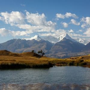 Un lago di montagna si trova in una valle erbosa di fronte a una lontana catena montuosa innevata sotto un cielo parzialmente nuvoloso.
