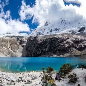 Un lago alpino turchese si trova ai piedi di una montagna aspra e innevata sotto un cielo blu con nuvole bianche.