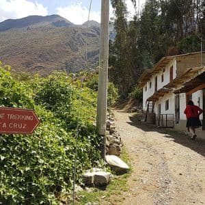 Un cartello rosso per il percorso di trekking Santa Cruz indica un sentiero sterrato che passa accanto a rustici edifici bianchi tra le montagne.