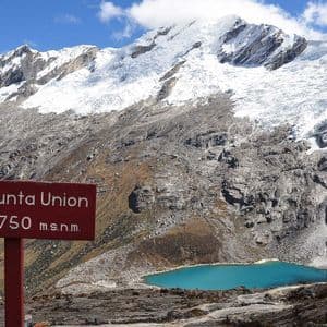 Un segnavia rosso si erge in un paesaggio roccioso con un lago alpino turchese sotto montagne innevate.