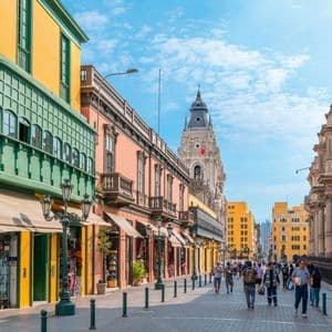 Gente passeggia lungo una soleggiata via pedonale fiancheggiata da colorati edifici in stile coloniale e una torre di chiesa in lontananza.