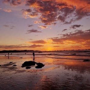 Silhouettes sur une plage mouillée à marée basse, avec un coucher de soleil coloré orange et violet se reflétant sur le sable et l'eau.