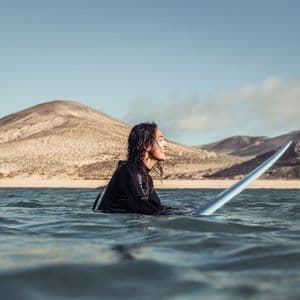 Una persona con i capelli bagnati in una muta nera sorride mentre è seduta su una tavola da surf nell'oceano, con colline sabbiose sullo sfondo.