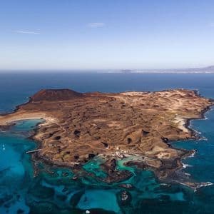 Una vista aerea di una piccola isola vulcanica arida circondata da acqua turchese e dal mare blu profondo, con barche a vela ancorate al largo.
