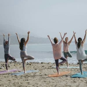 Cinque donne in viaggio di gruppo WeRoad che praticano la posizione yoga dell'albero su una spiaggia sabbiosa con vista sull'oceano.