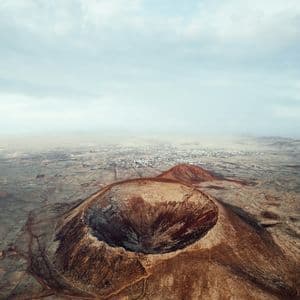 Una vista aerea di un grande cratere vulcanico in un paesaggio brullo e roccioso, con una piccola città visibile in lontananza sotto un cielo nuvoloso.