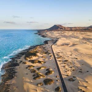 Una veduta aerea di una lunga strada che si snoda tra vaste dune di sabbia e una costa oceanica turchese, con montagne in lontananza.