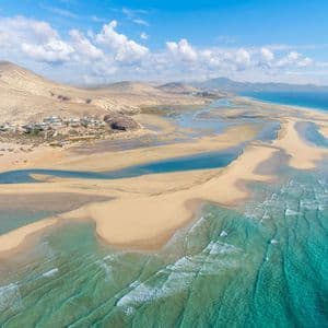 Vista aerea di una vasta costa sabbiosa che incontra le acque turchesi dell'oceano, con colline aride e un piccolo villaggio in lontananza.