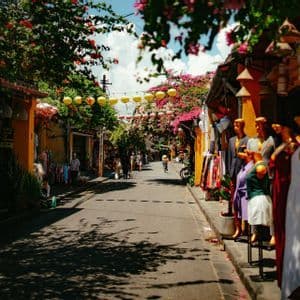 Una strada assolata fiancheggiata da negozi colorati, manichini e alberi fioriti, con lanterne gialle appese in alto.