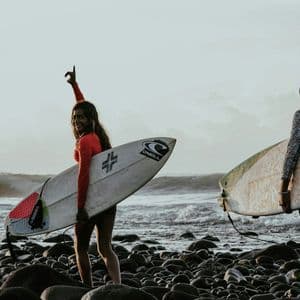 Due donne di un viaggio di gruppo WeRoad trasportano le loro tavole da surf su una spiaggia rocciosa, con le onde dell'oceano sullo sfondo.