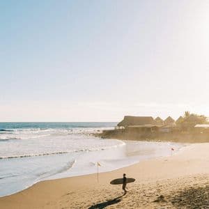 Un surfista in controluce cammina su una spiaggia sabbiosa con la sua tavola da surf, sullo sfondo l'oceano e le capanne balneari.
