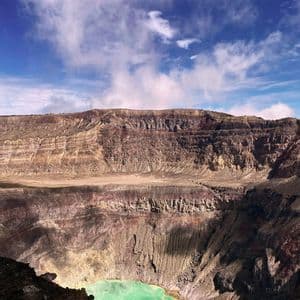 Una vista su un vasto cratere vulcanico con pareti rocciose stratificate e un lago turchese sul fondo, sotto un cielo blu con nuvole bianche.