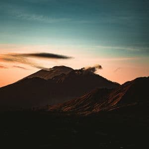 Una catena montuosa stagliata contro un tramonto dai colori vividi, con un pennacchio di fumo che si innalza da una delle vette.
