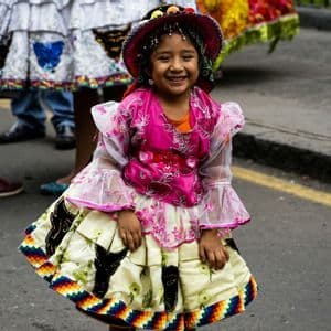Una giovane ragazza con un abito tradizionale colorato e un cappello sorride alla fotocamera, in piedi su una strada lastricata.